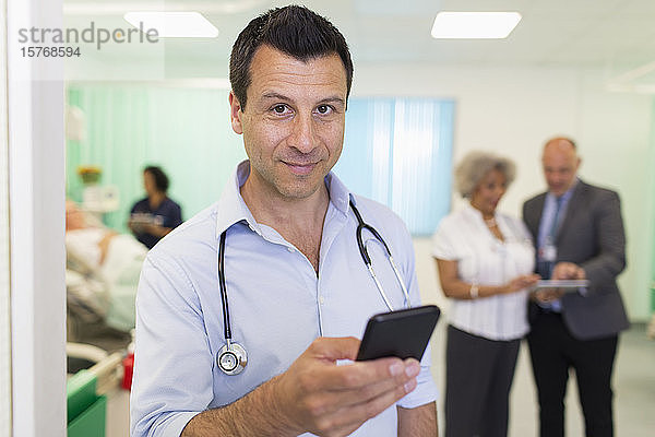 Porträt eines selbstbewussten männlichen Arztes  der ein Smartphone in einer Krankenstation benutzt