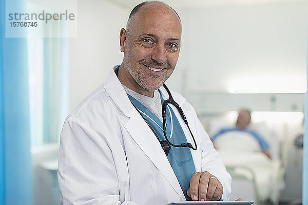 Porträt eines selbstbewussten  lächelnden männlichen Arztes  der ein digitales Tablet in einem Krankenhauszimmer benutzt