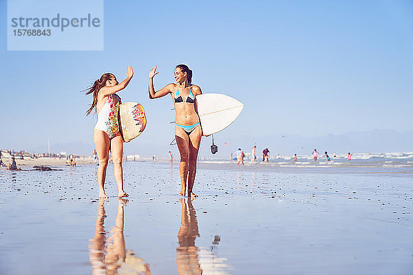 Glückliche junge Surferinnen  die sich am sonnigen Strand die Hände reichen