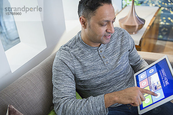 Mann stellt Smart-Home-Alarm über ein digitales Tablet auf dem Wohnzimmersofa ein