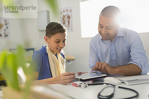 Männlicher Kinderarzt zeigt einem jungen Patienten in einer Arztpraxis ein digitales Tablet