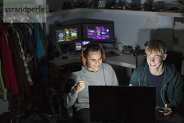 Aufgeregte Teenager mit Kopfhörern spielen ein Videospiel am Computer in einem dunklen Raum