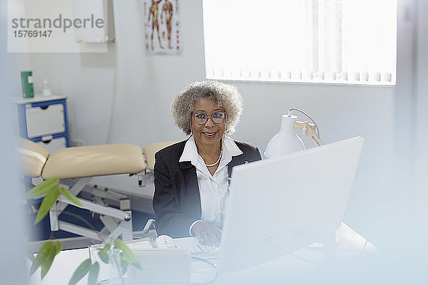 Porträt einer lächelnden  zuversichtlichen älteren Ärztin  die in einer Arztpraxis am Computer arbeitet
