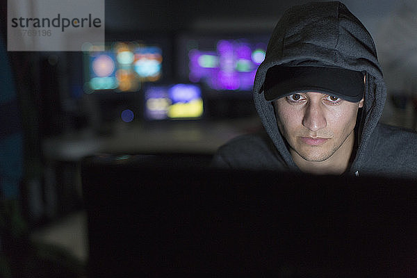 Männlicher Hacker im Kapuzenpulli  der einen Computer in einem dunklen Raum benutzt