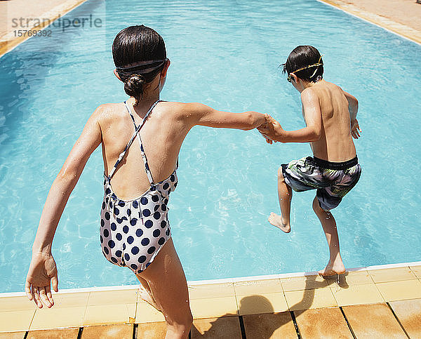 Junge und Mädchen halten sich an den Händen und springen in ein sonniges Sommerschwimmbad