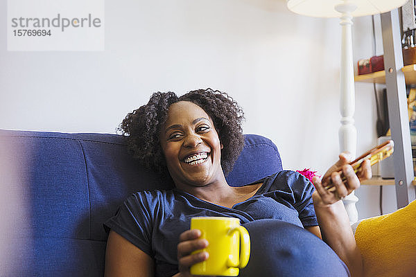 Lachende schwangere Frau mit Tee und Smartphone auf dem Sofa