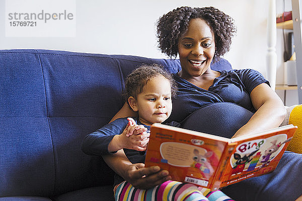 Schwangere Mutter liest ihrer Tochter auf dem Sofa ein Buch vor