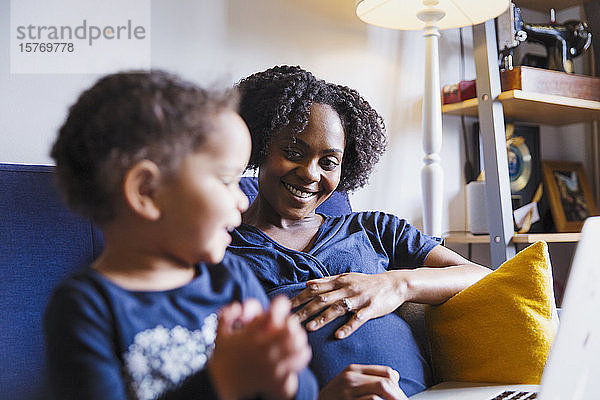 Glückliche schwangere Mutter und Tochter mit Laptop auf dem Sofa