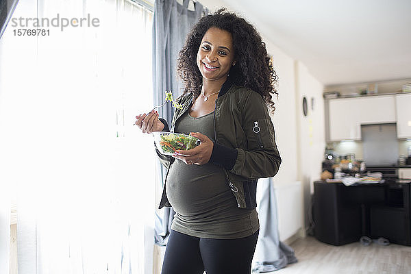 Porträt glückliche junge schwangere Frau isst Salat am Fenster