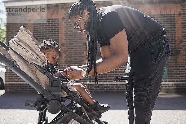 Vater  der sein Kleinkind im Kinderwagen auf dem sonnigen Bürgersteig festhält