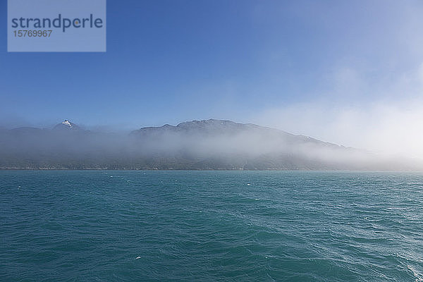 Nebelschwaden über Bergen und türkisblauem Meer Grönland