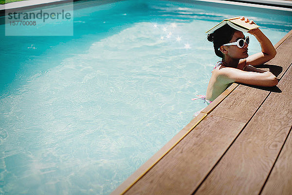 Frau hält Buch über Kopf in sonnigem Sommerschwimmbad