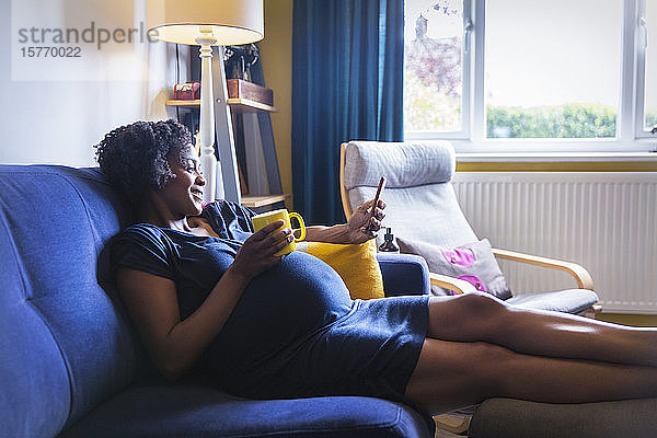 Glückliche schwangere Frau entspannt sich auf dem Sofa mit Tee und Smartphone