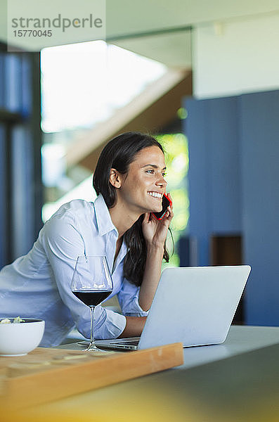Lächelnde Frau  die am Laptop mit einem Smartphone spricht und Rotwein trinkt