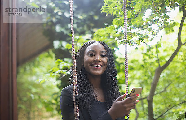 Porträt einer glücklichen jungen Frau  die auf einer Schaukel im Innenhof ihr Smartphone benutzt
