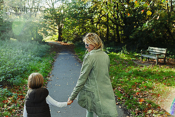 Glückliche Mutter und Sohn halten sich an den Händen und gehen in einem sonnigen Herbstpark spazieren