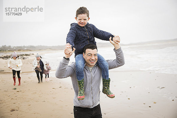 Porträt verspielter Vater trägt Sohn auf den Schultern am Winterstrand