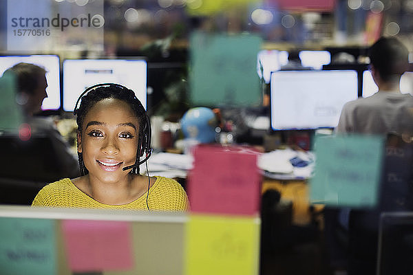 Porträt einer selbstbewussten  lächelnden Geschäftsfrau mit Headset  die am Computer hinter Haftnotizen im Büro arbeitet