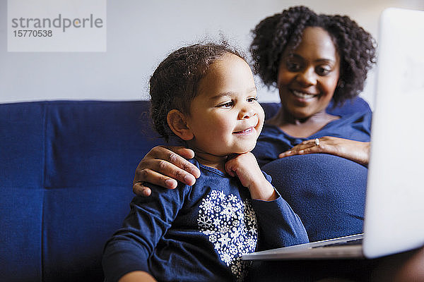 Glückliche schwangere Mutter und Tochter im Vorschulalter mit Laptop auf dem Sofa