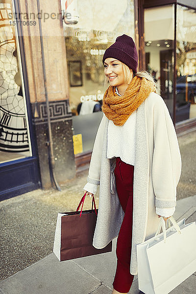 Junge Frau mit Strumpfmütze und Schal geht mit Einkaufstüten auf dem Bürgersteig