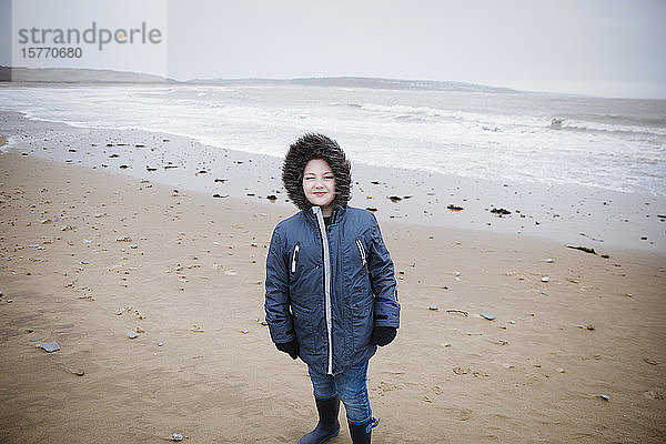 Porträt selbstbewusster Junge in Pelzkapuzenjacke am winterlichen Meeresstrand