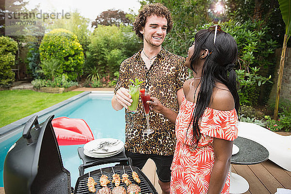 Glückliches junges Paar trinkt Cocktails beim Grillen am Pool