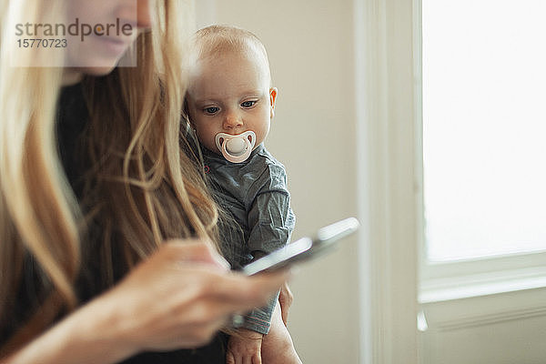 Neugieriges Baby-Mädchen mit Schnuller  das seine Mutter bei der Benutzung eines Smartphones beobachtet