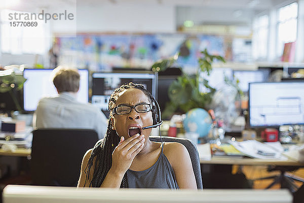 Müde Geschäftsfrau mit Headset gähnt am Computer im Großraumbüro