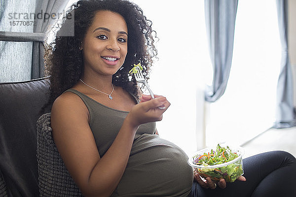 Porträt lächelnde junge schwangere Frau isst Salat auf Sofa