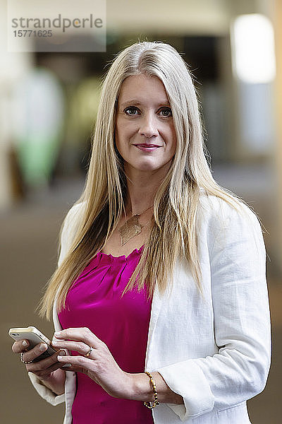 Eine professionelle reife Geschäftsfrau posiert für die Kamera in einem Flur  während sie ihr Smartphone auf Nachrichten überprüft: Edmonton  Alberta  Kanada