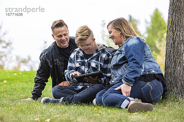 Ein junger Mann mit Down-Syndrom lernt mit seinem Vater und seiner Mutter ein neues Programm auf einem Tablet  während sie an einem warmen Herbstabend in einem Stadtpark die Gesellschaft des anderen genießen: Edmonton  Alberta  Kanada