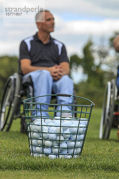 Ein Eimer mit Golfbällen im Vordergrund und ein Mann in einem Rollstuhl im Hintergrund