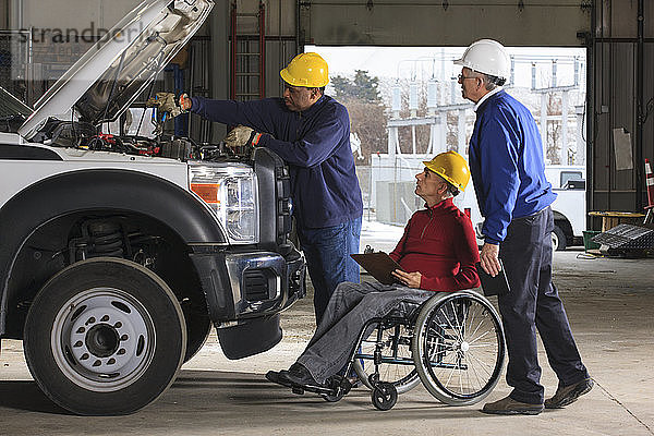 Kfz-Mechaniker bei der Arbeit an einem Lkw und Inspektoren  darunter ein Mann im Rollstuhl  die ihn bei der Arbeit beobachten