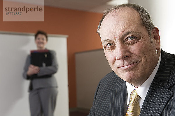 Blick auf einen lächelnden Geschäftsmann mit einer Geschäftsfrau im Hintergrund.