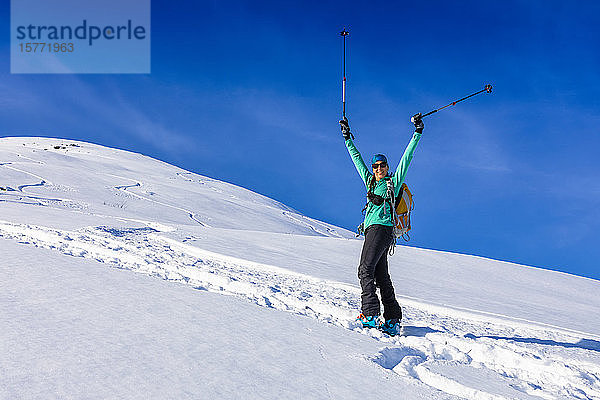 Frau beim Skilanglauf  die einen schönen Tag mit ihren Stöcken in der Luft feiert und mit AT-Skiern und Fellen in Hatcher's Pass  Alaska  Talkeetna Mountains  Alaska  Vereinigte Staaten von Amerika  den Berg hinaufklettert