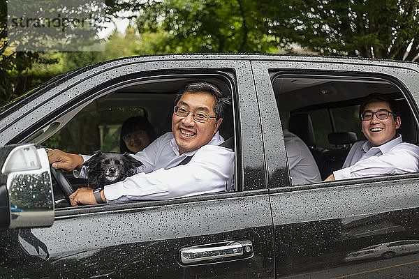 Familienfahrt im Auto mit dem Hund  der Vater am Steuer und die erwachsenen Kinder auf dem Rücksitz  die in die Kamera schauen und lächeln; Langley  British Columbia  Kanada