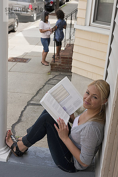 Eine junge Frau  die auf ihrer Veranda aus einem Lehrbuch liest und in die Kamera blickt  mit einer Mutter und ihrer Tochter im Hintergrund vor ihrem Haus