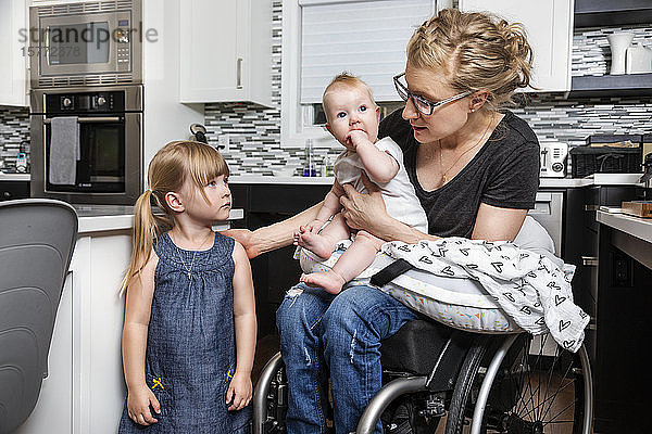 Eine querschnittsgelähmte Mutter im Rollstuhl spricht mit ihrer Tochter und hält ihr Baby auf dem Schoß  während sie in ihrer Küche arbeitet; Edmonton  Alberta  Kanada