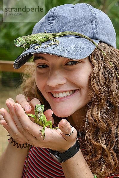 Porträt einer jungen erwachsenen Frau  die einen kleinen Leguan in der Hand hält und einen auf dem Schnabel ihrer Mütze sitzt  auf einer Leguanfarm; Roatan  Bay Islands  Honduras