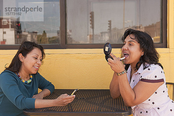 Zwei junge erwachsene Frauen sitzen draußen an einem Tisch  die eine trägt Lippenstift auf  die andere telefoniert mit ihrem Smartphone