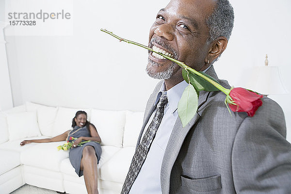 Porträt eines Mannes mittleren Alters  der eine Rosenblüte zwischen seinen Zähnen hält  mit einer lächelnden Frau mittleren Alters im Hintergrund