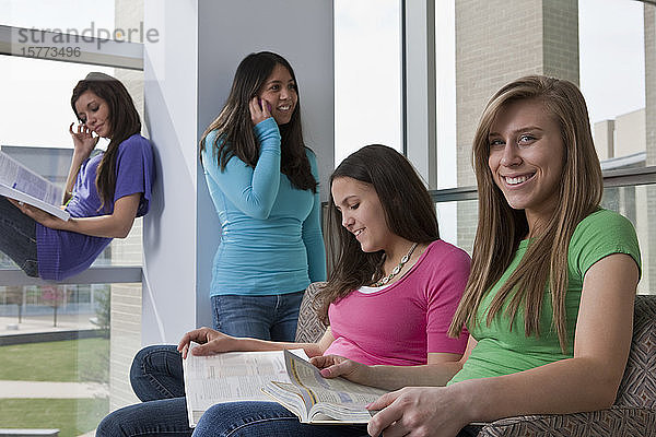 Vier Teenager-Mädchen auf dem Flur einer High School  eine mit einem Smartphone  die anderen blättern in Schulbüchern