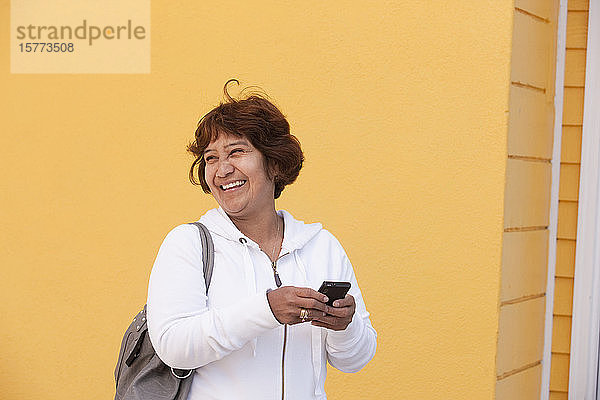 Eine ältere Frau steht an einer gelben Wand und hält ein Mobiltelefon in der Hand