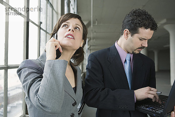 Geschäftsfrau im Gespräch über ein Mobiltelefon mit einem Geschäftsmann  der neben ihr einen Laptop benutzt
