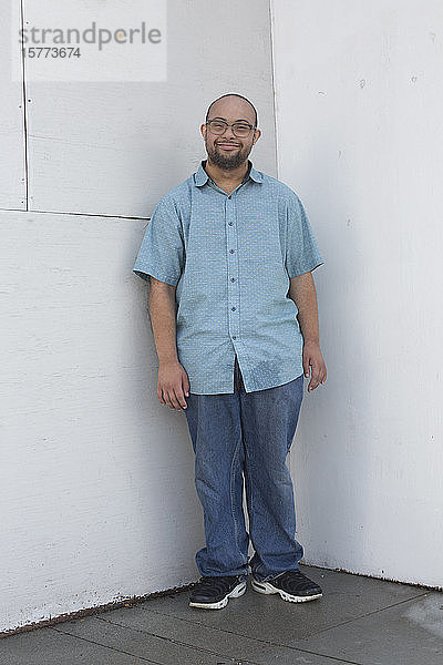 Porträt eines jungen Mannes mit Down-Syndrom  der in einer Ecke steht und in die Kamera schaut