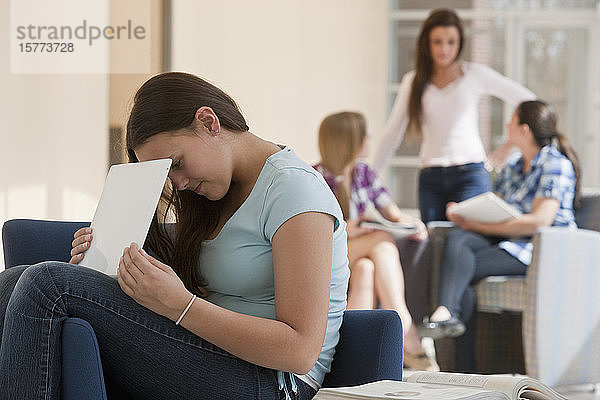 Teenager-Mädchen  das in der Schule einen Laptop benutzt und den Bildschirm vor den anderen versteckt  während zwei Schüler im Hintergrund auf Lehrbücher schauen