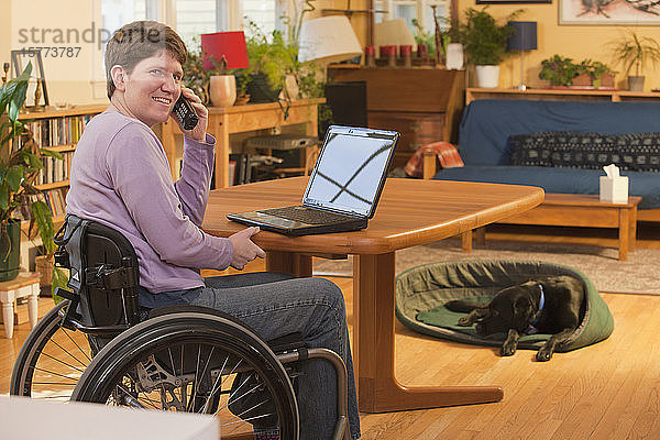 Behinderte Person im Rollstuhl  die zu Hause einen Laptop benutzt und mit ihrem Hund im Hintergrund telefoniert