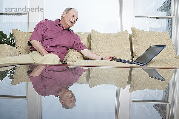 Älterer Mann sitzt auf einer Couch und schaut auf einen Laptop.