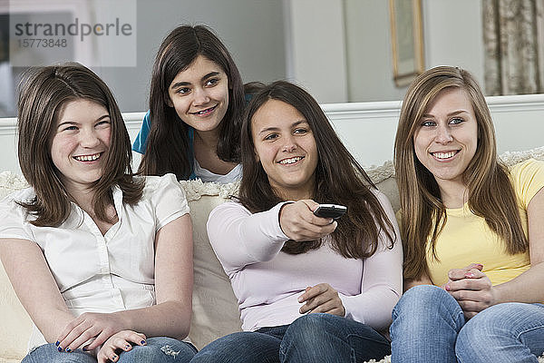 Vier Mädchen im Teenageralter  die zu Hause auf einer Couch sitzen  fernsehen und mit der Fernbedienung den Kanal wechseln