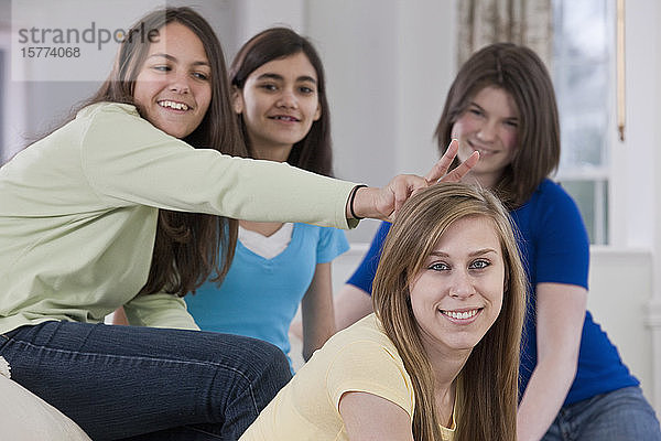 Porträt von vier Mädchen im Teenageralter  die zu Hause auf einer Couch sitzen  wobei ein Mädchen ihrer Freundin aus Dummheit Hasenohren hinter den Kopf steckt